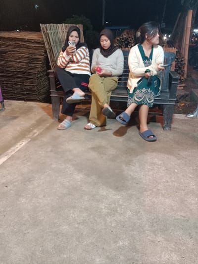 Three women are seated on a dark-colored bench at night in Sekopek Wetan, Indonesia, on a light grey concrete surface. The woman on the left wears a black hijab, a white and brown striped long-sleeved shirt, black pants, and light blue slide sandals, looking at a white smartphone held in both hands. Her left leg is crossed over her right. The middle woman wears a dark hijab, a light grey long-sleeved top, olive green pants, and white slide sandals, holding a small red object with both hands. Her legs are crossed at the ankles. The woman on the right wears a dark green patterned dress with a white long-sleeved cardigan and dark blue slide sandals. Her dark hair is styled up, and she is looking to her right, away from the camera. Behind the bench, to the left, is a stack of dark, woven material, possibly dried bamboo mats. To the right, behind the bench, are stacked bamboo poles. Distant lights are visible in the dark background.