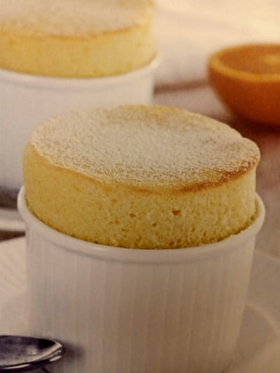 A close-up shot captures a fluffy, golden-brown soufflé dusted with powdered sugar, served in a white ramekin. The soufflé is moist and airy, with a slightly caramelized top. In the background, a sliced orange in its peel sits in a blurred orange bowl, suggesting a citrus flavor profile. The setting appears to be a restaurant in Rumaithiya, Kuwait.