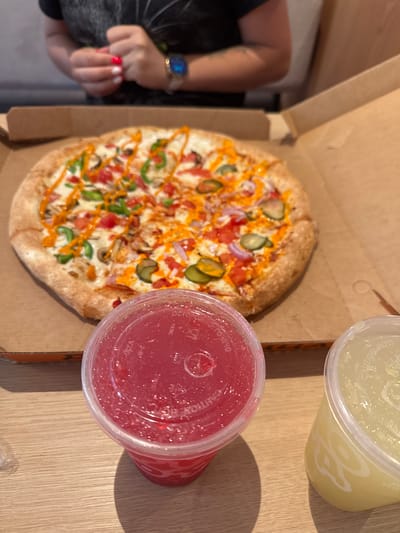 In Koltsovo, Russia, a pizza is presented in its cardboard box, situated on a wooden surface. The pizza is topped with tomatoes, green peppers, red onions, mushrooms, and a drizzle of orange sauce. In the foreground, two plastic cups are visible. The left cup contains a red beverage, and the right cup holds a yellow beverage. In the background, a person wearing a black t-shirt is partially visible, with their hands clasped and a watch on their left wrist. The environment appears to be an indoor setting, likely a restaurant.