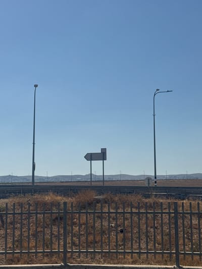 The image depicts a roadside scene with a clear blue sky and a dry, barren landscape. In the foreground, a metal fence runs horizontally across the bottom of the frame. Behind the fence, dry brush and vegetation are visible. Two tall streetlights stand on either side of the frame, with a directional signpost located between them. The signpost has a white rectangular sign with a black arrow pointing to the left. In the background, a series of hills and a distant horizon are visible, with scattered structures and more streetlights. The overall impression is one of a semi-arid environment, possibly along a highway or road in the Bnei Shimon Regional Council, Israel, characterized by its open spaces and sparse vegetation.