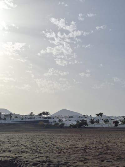 This is an outdoor, eye-level, full shot taken in La Concha, Spain. The foreground is dominated by a sandy beach with a textured surface. In the midground, a low white wall runs horizontally across the frame, separating the beach from a cluster of white buildings and palm trees. The buildings are low-rise, with dark windows and some red-tiled roofs. Several palm trees are scattered throughout the scene, some taller and more slender than others. In the background, rolling hills or mountains with smooth, rounded peaks are visible, partially obscured by the buildings and trees. The sky is a pale blue with scattered, wispy white clouds. Sunlight streams in from the left, creating a slightly hazy effect and casting subtle rays across the scene. There are no people or animals clearly visible in the frame.