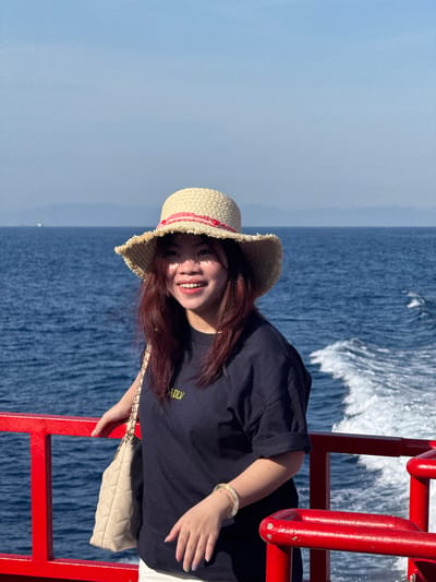 A young East Asian woman wearing a straw hat and a dark blue t-shirt is standing on the deck of a boat, with the sea and sky as the background. The boat has red railings. She is holding a quilted beige handbag on her left shoulder and her right hand is resting on the railing. She is smiling and looking to her left. The sea is a deep blue with white waves and foam trailing behind the boat. In the distance, a hazy mountain range is visible under a clear blue sky. This scene appears to be on the waters near Dong Son city, Vietnam.