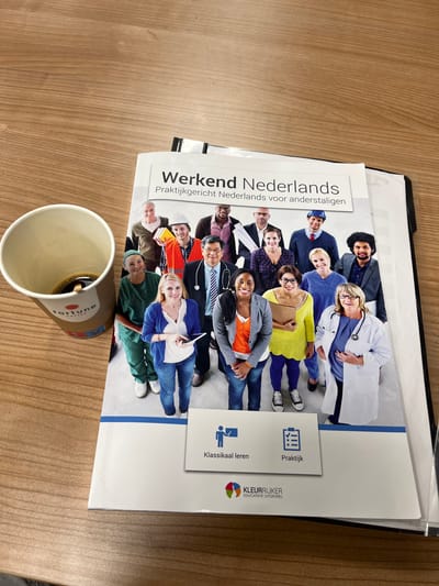 An eye-level, medium shot shows a book titled "Werkend Nederlands" resting on a wooden table next to a disposable coffee cup. The book features a collage of diverse individuals in various professional attire, suggesting a theme of employment and integration. The coffee cup, branded with "fortune COFFEE," contains a dark liquid. The book is slightly angled, revealing a binder behind it. The wooden surface appears to be a desk or table. The background is out of focus. There is no visible information to indicate the location in the city of Oldenzaal, Netherlands.
