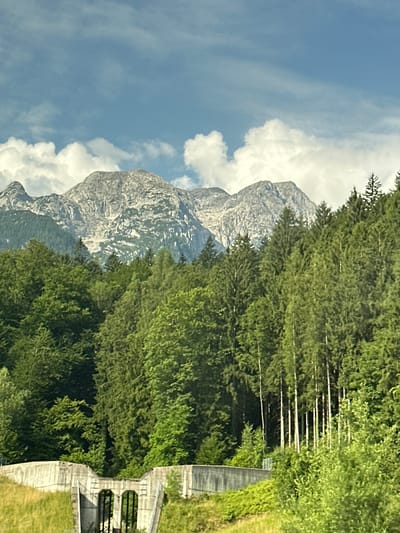 The image depicts a scenic view of the Kuchl, Austria, area, focusing on a mountainous landscape. In the foreground, a concrete structure, possibly a bridge or an underpass, is visible. This structure has arched openings and is partially covered in vegetation on its sides. A grassy slope leads up to the structure. Behind this, a dense forest of evergreen and deciduous trees covers the hillside, with a clear distinction between the darker evergreens and lighter deciduous trees. Towering above the forest are rugged, rocky mountains with some patches of snow or lighter rock formations. The sky above is a clear blue with scattered white, wispy clouds. The overall impression is one of a natural, mountainous environment with some human infrastructure integrated into the landscape.