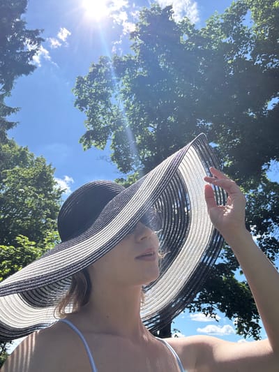 A young woman, partially obscured by a large, wide-brimmed hat with black and white stripes, stands outdoors under a bright, sunny sky. Her right hand is raised, holding the edge of the hat. The hat casts a shadow over her face, though her lips and chin are visible. She wears a light blue strap, possibly from a swimsuit or dress. In the background, lush green trees with visible foliage frame the sky, which is a vivid blue with scattered white clouds and intense sunrays. The image appears to have been taken on a warm, clear day. The location is identified as Lendži, Latvia.