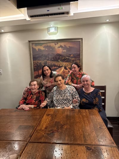 Five women of varying ages pose for a photograph indoors, likely in a restaurant. The setting is suggested by the large wooden table occupying much of the foreground. Three women sit at the table while two others stand behind them.

The woman seated on the left wears a dark floral print blouse and a wristwatch. Next to her, another woman is dressed in a light-colored patterned blouse, also with a wristwatch. On the right side of the table, the third woman has on a dark floral dress with long sleeves.

Behind them, the woman on the left is wearing a light floral blouse. The woman on the right, standing behind the seated woman in the dark floral dress, wears a brightly colored floral top.

A framed painting depicting a landscape hangs on the wall behind the women. Above the painting is an air conditioning unit and a light fixture.

The location is assumed to be in Tijuana, Mexico, but there is no forensic evidence of such in the photo.