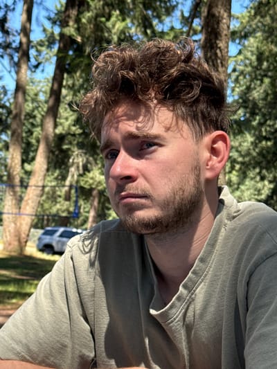 The image is a close-up, outdoor portrait of a young Caucasian male with medium-length, curly brown hair and a light beard. He is wearing a light green t-shirt and is looking off to his left with a pensive expression. The background is blurred but shows a park-like setting with tall trees and foliage under a bright blue sky. A volleyball net and a white SUV are visible in the background. The lighting suggests it is daytime.
