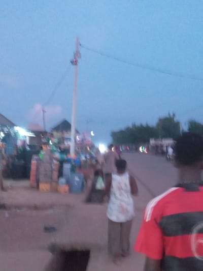 The image, taken in Eka, Nigeria, depicts a street scene at dusk. The sky is a light blue. A utility pole rises on the left side of the frame, with power lines extending from it. Streetlights are visible along the right side of the image, blurred due to the low lighting and motion blur. People are present on the right side of the image. One individual wears a shirt with red and black stripes. Another person is wearing a white dress. Stalls or shops are seen further down the street, illuminated by lights. The scene is generally blurry, suggesting a moving camera or low light conditions.