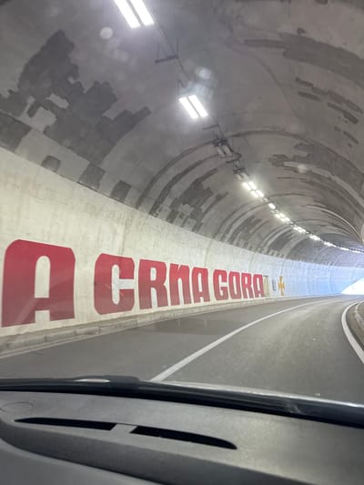 The image shows the interior of a tunnel, likely the Sozina Tunnel, located in Bukovica, Montenegro. Large red lettering on the left wall spells out "CRNA GORA", the Montenegrin name for Montenegro. To the right of the text, there is a yellow cross with four equal arms. The tunnel has a concrete ceiling with visible repairs and lighting fixtures. The road within the tunnel is asphalt with white lane markings. The perspective suggests the photo was taken from the driver's seat of a vehicle. The overall scene is industrial and utilitarian, characteristic of a road tunnel.