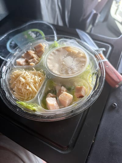 A clear plastic container holds a salad with grilled chicken pieces, shredded cheese, and lettuce. A small container of dressing sits in the center of the salad. The container is placed on a black surface. A knife with a wooden handle is positioned to the right of the salad container. The background is out of focus and appears to be an indoor setting. The image is taken from a slightly elevated angle, looking down at the salad. There are no people visible in the image. The location is not specified in the image or provided.