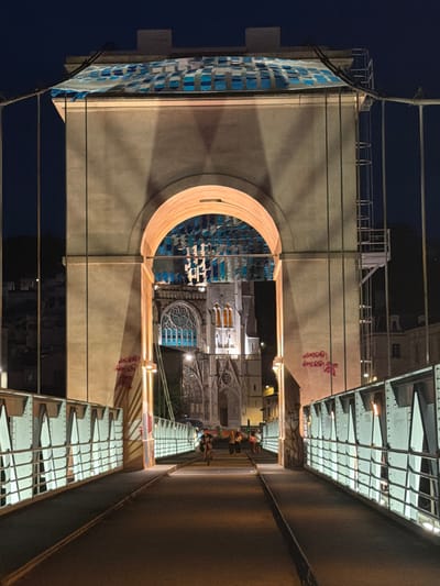 The image shows a nighttime view of the Pont du Marché in Vienne, France. The bridge, which is illuminated by artificial lights, features a large archway that frames a view of a Gothic cathedral in the background. The cathedral has intricate stonework and stained-glass windows, which are also lit from within. On the bridge, a person is riding a bicycle and several other individuals are walking. The bridge is lined with metal railings that are also lit, casting a soft glow on the walkway. The night sky is dark, with no stars visible. The overall atmosphere is one of urban tranquility, with the illuminated bridge and cathedral creating a striking contrast against the dark night.