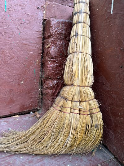 A straw broom leans against a dark reddish-brown wooden wall and a rough, dark red brick corner. The broom's handle is bound with dark string in several places, creating a cylindrical shape. The bristles of the broom are made of straw and are fanned out on the ground. The ground appears to be a weathered, reddish-brown surface with some lighter paint flecks. The overall scene suggests an outdoor or semi-outdoor environment. The location is Altayskiye Zori, Russia.