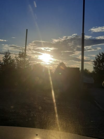 This media file captures a sunset scene in Shelekhov, Russia. The main subjects are a clear blue sky with scattered, wispy clouds illuminated by the setting sun, and silhouetted trees and a tall pole in the foreground. The sun is a bright, blown-out white orb low in the sky, casting a warm, golden light and lens flares across the image. The scene appears to be viewed from inside a vehicle, as indicated by the reflection on the right side of the frame, which shows a surface with some dirt or water spots and a subtle sheen. The overall mood is peaceful and serene, with the natural beauty of the sunset dominating the composition. The time of day is clearly sunset due to the quality of light and the position of the sun. There are no people or discernible text visible in the image.