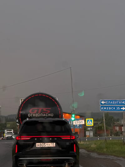 Here is a detailed description of the image:

**Location:** The image appears to have been taken in Minderevo, Russia, based on the road sign indicating directions to "ЕЛАБУГА 142" and "ИЖЕВСК 35."

**Scene:** The image shows the rear of a black EXEED vehicle stopped at what appears to be a traffic light. A tank labeled "ОГНЕОПАСНО" (which means "Flammable" in Russian) is attached to the vehicle.

**Vehicle Details:**
*   The license plate of the black EXEED vehicle reads "M805 TY 13."
*   The tank behind the vehicle has the letters "GTS" and the word "ОГНЕОПАСНО" written on it, circled by a dashed red outline.

**Traffic Signals and Signage:**
*   The traffic signal is green.
*   A "СТОП" (Stop) sign and a pedestrian crossing sign are visible in the background.

**Environment:**
*   The weather appears to be overcast or rainy.
*   There is greenery visible in the background along the roadside.
*   Several other vehicles are visible in the distance.