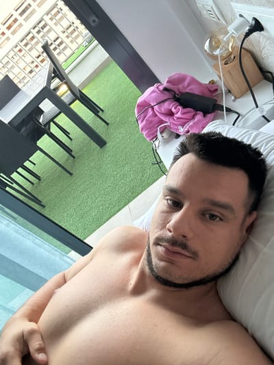 A man is lying on a pillow in a bedroom. He is shirtless and has a beard. In the background, there is a table and chairs on a patio, and a building with balconies. There are also some electrical outlets and chargers on a nightstand. The image appears to have been taken indoors, looking out towards a patio area. The overall scene suggests a domestic setting, possibly a hotel room or a residence, in Roquetas de Mar, Spain.