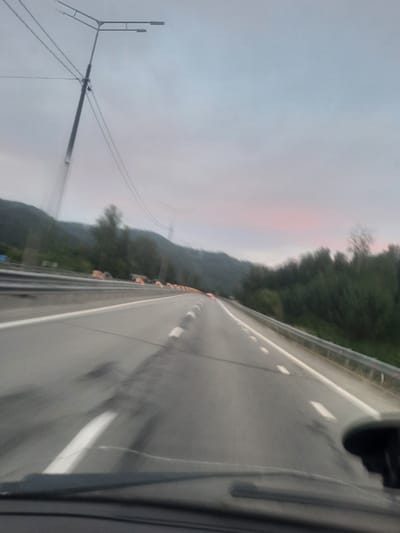 The image depicts a view from a moving vehicle on a highway, likely in Rybalka, Russia. The road, with white lane markings and reflective bollards, curves to the right. On the left side of the road, a line of trees forms a dense forest that ascends a mountainside. The sky is overcast, with a faint pinkish hue suggesting either sunrise or sunset. In the distance, a large, lattice-like structure, possibly a transmission tower, is visible against the sky. Another pole with multiple lights is also present on the left side of the road. The overall mood is serene and perhaps a bit melancholic due to the dim lighting and muted colors. The motion blur indicates that the photograph was taken while the vehicle was in motion. There is no visible text in the image.