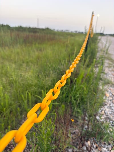 A close-up, low-angle, outdoor shot depicts a bright yellow plastic chain fence stretching diagonally from the lower left corner of the frame towards the upper right. The chain links are large and hollow, forming a repeating pattern against a background of green grass and a gravelly path. In the distance, the chain fence continues, bordered by a light pole with an illuminated lamp. The sky above is a pale, overcast gray, suggesting either dawn or dusk. The environment appears to be a rural or undeveloped area, possibly on the outskirts of Cardwell, United States, with tall weeds and sparse vegetation. There are no people or animals visible in the image.