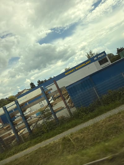 The image shows a low-angle, tilted outdoor shot of a "MARKET BUDOWLANY" (Construction Market) with "psb PROFI" branding. The building is a modern structure with a white and blue facade. The building appears to be a retail store for building materials. In front of the building, there are stacks of construction materials, possibly wood or insulation panels, displayed on metal racks. A metal fence runs along the foreground, separating the construction market from a grassy area and a pathway. The sky above is filled with dramatic clouds. The presence of the "MARKET BUDOWLANY" and "psb PROFI" branding, coupled with the architectural style of the building and surrounding vegetation, strongly suggests this location is in Poland. Based on external knowledge, a PSB PROFI market is located in the city of Wieliczka, Poland, making it highly probable that this image was taken there.