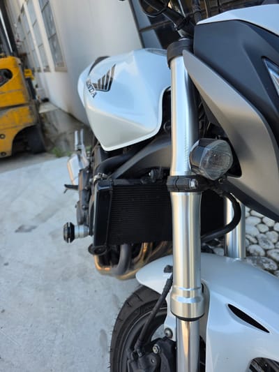 The image shows a close-up, low-angle view of the front section of a white Honda motorcycle parked on a concrete surface. The motorcycle's front forks, radiator, a portion of the fuel tank with the Honda logo, and the front fender are visible. In the background, a yellow forklift and a white building with barred windows are present. The environment suggests an outdoor urban setting, possibly a workshop or storage area, in Stanghella, Italy. No people are present in the image.