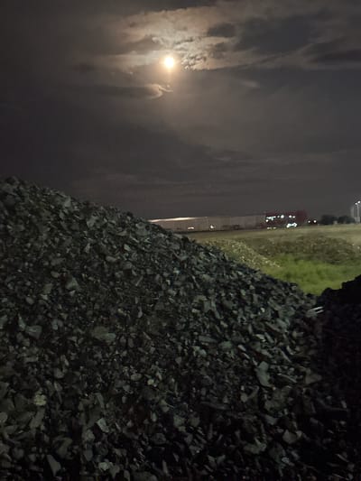 This nighttime photograph, taken in Arad, Romania, captures a landscape characterized by industrial elements and natural features under a cloudy sky. The image is oriented vertically, with the bottom half dominated by a large, dark pile of angular rocks, resembling gravel or railway ballast, which fills the foreground and extends towards the right side of the frame.
Above the rocky foreground, a strip of ground is visible, partly covered in sparse, dark green vegetation. Further in the background, a series of buildings or industrial structures are illuminated by numerous small, bright artificial lights, suggesting an active urban or industrial area in the distance.
The upper half and left side of the image are occupied by a cloudy night sky. A prominent, bright moon shines through the clouds, its light diffused and illuminating the surrounding cloud formations with a soft glow. The overall impression is one of a quiet, atmospheric night scene with no human activity or figures present. There is no visible text in the image.