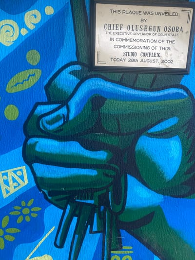 This image is a close-up photograph of a plaque mounted on a brightly colored mural. The plaque is rectangular with a dark frame and has text inscribed on it. The text reads: "THIS PLAQUE WAS UNVEILED BY CHIEF OLUSEGUN SOBA THE EXECUTIVE GOVERNOR OF OGUN STATE IN COMMEMORATION OF THE COMMISSIONING OF THIS STUDIO COMPLEX TODAY 28th AUGUST, 2002." The mural behind the plaque features a stylized depiction of a clenched fist in shades of blue and green, with various abstract patterns and shapes in yellow and blue. The mural is textured, suggesting it is painted on a wall. The location is identified as the city of Ojere, Nigeria.