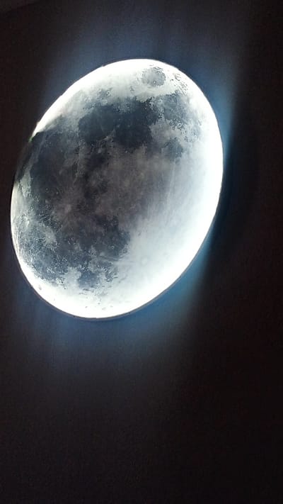 A large, circular, illuminated object resembling the moon is affixed to a dark wall. The object displays realistic lunar textures and craters. A bright, white light emanates from behind the object, creating a halo effect around its circumference. The majority of the image is taken up by the dark wall, with the illuminated moon occupying the upper left quadrant. There is no indication of the city of Choktal, Kyrgyzstan, or any people in the image.