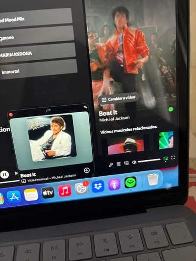 The image displays a close-up view of a laptop screen, which is showing a media playback interface, likely a music or video streaming application. The left side of the screen features a dark gray sidebar with a list of song or playlist titles: "Good Mood Mix," "Simone," "MARIMANDONA," and "Immortal."
The right side of the screen shows album art for Michael Jackson's "Thriller" album, with the song title "Beat It" and artist "Michael Jackson" displayed below. A "Change to video" button is visible. Further down, a section labeled "Related music videos" shows thumbnail images of music videos. A progress bar and playback controls are visible at the bottom of this section.
The bottom of the laptop screen displays a dock with various application icons, including a blue Apple logo, a notepad icon, an Apple TV icon, a red music note icon, a gear icon with a notification badge, and a blue tile icon. Other icons visible include Dropbox, Podcasts, Spotify, and a trash can.
The laptop keyboard is partially visible at the bottom of the image, with several function keys labeled. The overall environment appears to be indoors, with a portion of a red and white patterned surface visible in the lower right corner. There is no identifiable information about the specific location within "Urbanización Encinares De Alcolea, Spain."