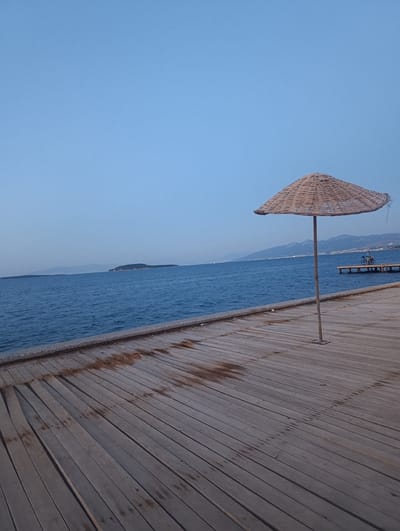 A serene scene unfolds on a wooden pier under a clear, pale blue sky, suggesting either early morning or late evening. The vast, calm blue sea stretches out to the horizon, where a small, dark island is visible in the distance, flanked by hazy mountains. The wooden planks of the pier, weathered and displaying some dark, damp patches, lead the eye towards the water.

Dominating the foreground is a simple, woven straw umbrella, its natural fibers creating a textured canopy supported by a thin metal pole. To the right, further along the pier, a small wooden structure with steps descends into the water. Perched on this structure is a bicycle, and a person can be seen standing beside it, looking out towards the sea. The overall atmosphere is peaceful and tranquil, evoking a sense of quiet contemplation or the end of a day's activity. The clear sky and calm water indicate fair weather. The location is Urla, Turkey, a coastal area known for its natural beauty. No visible text is present in the image.