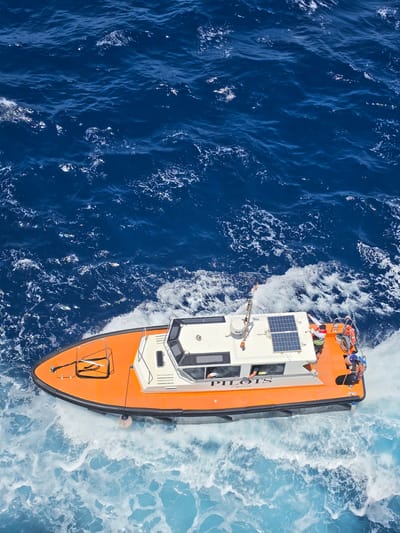 This is an aerial shot of an orange and white pilot boat moving through deep blue water, with white foam churned up by its wake. The boat has a cabin structure on its deck, equipped with solar panels. A person wearing a life vest is visible on the deck. The word "PILOTS" is emblazoned on the side of the cabin in large black letters. The scene appears to be set in a nautical environment, likely offshore, given the presence of the pilot boat. The water is a vibrant blue, suggesting a clear day, possibly daytime, with sunlight reflecting off the surface. The wake indicates the boat is in motion, actively navigating the water. The overall impression is one of maritime activity and operational readiness.