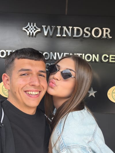 A man and a woman pose for a photo in front of the Windsor Convention Center in Bayrampaşa, Turkey. The man is in the foreground, smiling broadly, and wearing a black long-sleeved shirt. The woman is behind him, pouting, wearing sunglasses, and a denim jacket. The building sign behind them says "WINDSOR CONVENTION CENTER" vertically, with a round logo to the right.