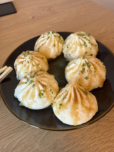 A close-up, overhead shot shows a circular, dark plate filled with six pan-fried dumplings. The dumplings are arranged in a circular pattern around the plate, with one in the center slightly obscured by the others. They have a golden-brown bottom from pan-frying and a white, pleated top. Each dumpling is drizzled with a light brown sauce and sprinkled with chopped green onions and black pepper. The plate sits on a light brown wooden table. Two chopsticks, with some visible text on them, are positioned near the bottom of the plate, slightly angled. The lighting suggests daytime. The scene is a food presentation, likely in a restaurant or home setting in Damme, Belgium.
