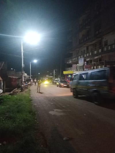 The image depicts a nighttime street scene in Kahuro, Kenya, illuminated by artificial light sources. A bright overhead street light on a pole dominates the mid-left foreground, casting strong illumination on a dirt or unpaved road. To the immediate left of the road, a strip of green grass and dirt borders several small, single-story structures with corrugated metal roofing, one displaying a partially visible "Airtel" sign. Several motorcycles are parked near these structures.
On the roadway, two individuals are visible in the mid-ground. One person stands prominently in the center-left, wearing light-colored clothing, facing towards the viewer. Another person, also in light attire, stands further down the road near the parked motorcycles. Vehicular traffic is present: a dark-colored van with a yellow stripe is in motion on the right side of the road, partially cut off by the frame, traveling towards the viewer. Behind it, a dark sedan with illuminated headlights is visible, facing away. Additional vehicle headlights are discernible further down the street.
The right side of the scene is dominated by a multi-story building, appearing to be at least five floors, featuring multiple balconies. Signs are visible on this building, including "HUSTLER KINYOOZI" and a partially visible "Optica" sign. A bright yellow kiosk or small structure is located at the base of this building. The sky is dark, indicating night conditions.
