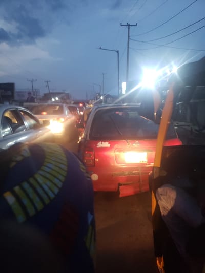 The image shows a traffic jam at night in Ida, Nigeria. Several vehicles, including a red van with its lights on, are congested on a road. The sky is visible in the background, suggesting it's either dusk or dawn. Utility poles and wires are visible along the roadside. The image is slightly blurred and taken at an angle. There is a person sitting in the driver’s seat.