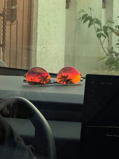 A pair of sunglasses with red lenses reflects a palm tree, indicating a warm climate. The sunglasses are hanging from a car's dashboard or console. The background visible through the window includes foliage and what appears to be a building exterior. The presence of a steering wheel and other interior car elements is noted. It is impossible to confirm the location of the scene as Oacalco, Mexico.