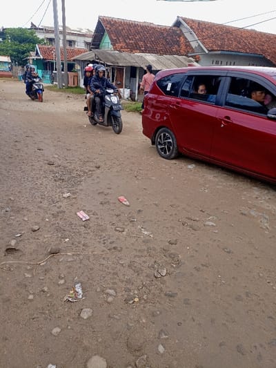 The image shows a street in Pameungpeuk, Indonesia, located at the coordinates -7.668 latitude and 107.694 longitude.  The scene is a dusty, unpaved road, with a red car parked on the right side and a motorbike parked to the left.  The car is facing away from the camera with the rear window open.  Three people are seated in the back of the car.  The motorbike has two people riding on it.  In the foreground, there are three pieces of pink trash on the ground.