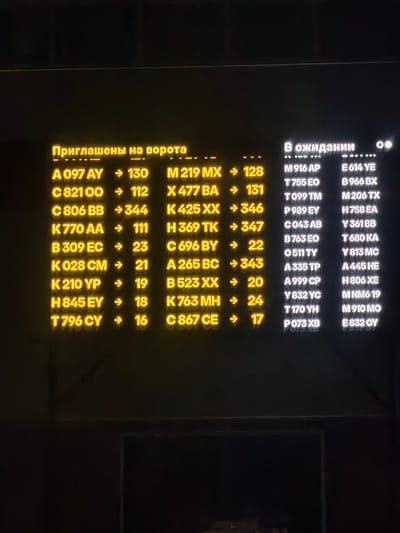 The image shows a digital display board with yellow and white text on a black background. The text is in Russian and English. The top of the display board has the Russian text "Приглашены на ворота", which translates to "Boarding at the gates". Below this, there are lists of codes and numbers, some with arrows, suggesting flight or gate information.
To the right of the yellow text, there is a section with white text that reads "В ожидании", which translates to "Waiting". This section also lists codes and numbers.
Based on the visual cues of a digital display board commonly found in airports, and the Russian text, this image likely depicts a departures or arrivals board at an airport. Without further information or context, it is not possible to definitively determine the specific location within Sluchainyi, Russia. However, such displays are characteristic of airport terminals.
The subjects are the text on the display board. The objects are the digital display board itself. The environment is likely an indoor public space, possibly an airport terminal. There are no people directly visible in the image, only the information presented on the screen. The lighting appears to be dim, with the display board being the primary light source in its immediate vicinity.
