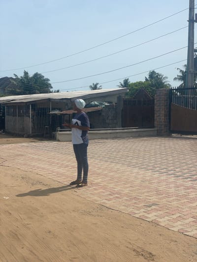 A person wearing a white cap, a dark and white shirt, and dark pants stands near a building and a red brick patterned walkway in Ikoga, Nigeria. Overhead power lines are visible. The person’s shadow is cast on the sandy ground adjacent to the walkway.