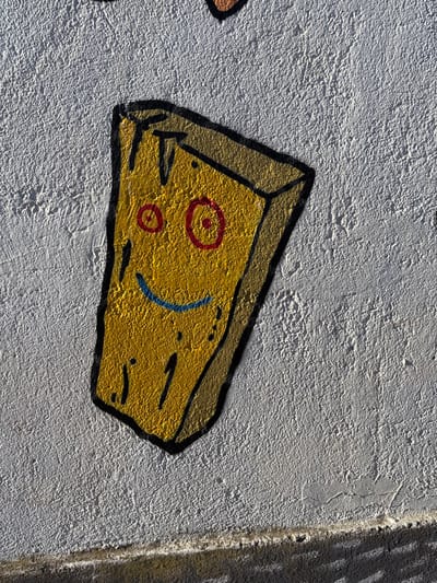 The image displays a detailed view of a white, textured wall located in Tomé, Chile, adorned with a piece of graffiti art. The central subject is a stylized, yellow, rectangular, wedge-shaped object, outlined in thick black and shaded with brown, visually resembling a block of cheese or wood. This object features a cartoon-like face: two prominent red circular eyes with smaller red pupils, and a horizontal blue curved line forming a smile. The yellow surface of the object also contains several small black vertical lines and dots. The wall, consistent with stucco or concrete, exhibits a rough, granular texture and minor surface imperfections. A dark, horizontal line or architectural ledge runs across the bottom section of the frame. The scene is illuminated by natural light, highlighting the textures and colors of the art and the wall.