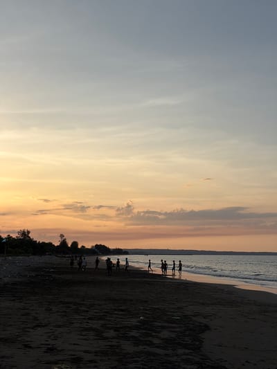 The image depicts a beach scene in Pasi Rawa, Indonesia, during sunset. The sky is a gradient of soft blues and oranges, with scattered clouds. The foreground is a dark, sandy beach with a line of trees and vegetation along the horizon. In the middle ground, silhouetted figures of several people, possibly children, are engaged in an activity resembling playing soccer on the wet sand near the water's edge. The ocean is calm, with gentle waves lapping at the shore. The overall atmosphere is serene and tranquil.