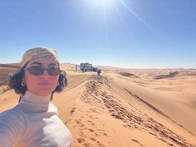 The image depicts a desert landscape, likely Erg Chebbi near Rissani, Morocco, under a clear, bright blue sky with a prominent sun flare in the upper left. In the foreground, a female subject is positioned on the crest of a large, undulating sand dune, taking a selfie. She wears dark, oval-shaped sunglasses, a patterned headscarf in shades of yellow, green, and pink, a light beige ribbed turtleneck top, and large, light-colored circular hoop earrings. Her dark, curly hair is visible around her face.

In the mid-ground, further along the dune crest to the right of the primary subject, a white 4x4 off-road vehicle, resembling a Land Rover Defender, is parked. Two additional figures are visible near the vehicle; one is standing next to the driver's side, and another is partially obscured by a dune, appearing to be crouched or positioned low to the ground.

The environment consists of numerous vast sand dunes of varying sizes, exhibiting shades of orange and tan, with visible footprints and tire tracks on their surfaces. Shadows cast by the dunes indicate bright sunlight. The background extends into a hazy horizon dominated by more distant sand dunes.