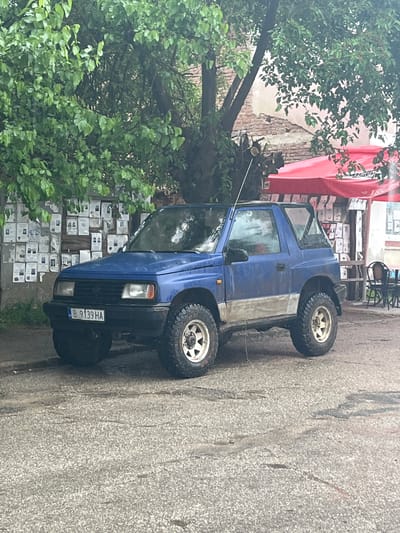 The image, taken in Kramolin, Bulgaria, shows a blue off-road modified compact SUV parked on a paved surface during the daytime. The vehicle, possibly a Suzuki Vitara or similar, features large, knobby tires mounted on white multi-spoke rims, indicating its off-road capability. The body of the SUV appears weathered or dusty, with a distinct two-tone effect of blue on the upper half and a lighter, greyish-white on the lower side.
Dense green foliage from several trees frames the left side and foreground of the image, with a thick tree trunk visible behind the car. To the right of the vehicle, a building or structure is partially visible, featuring a bright red awning or tarp, possibly belonging to a cafe or shop. Below this awning, a wall or fence is almost entirely covered with numerous white rectangular papers, which appear to be advertisements, notices, or bulletins, creating a visually busy backdrop. In the background under the awning, stacked items (possibly boxes) and some dark chairs (suggesting outdoor seating) can be seen.
The car is parked on a grey, roughly textured paved street. The lighting is diffused, indicating either an overcast day or sunlight filtered through the surrounding trees. No human activity is visible, and the scene presents a static street view. The license plate on the front of the SUV clearly displays "B 9139 HA," with the "B" prefix signifying registration in Sofia, Bulgaria.