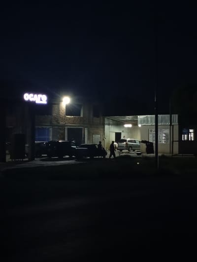 The image depicts a nighttime scene in an urban setting, likely a commercial area. In the background, a building with exposed brickwork and large window openings is visible. A prominent illuminated sign with the text "OGATO" in large, purple, stylized letters is mounted on the left side of the building. A bright overhead light shines from the upper right of the building. In front of the building, several vehicles are parked, including sedans and what appears to be an SUV. Two individuals are visible near the vehicles: one is standing in the foreground, facing away from the camera, and another is walking towards the right of the frame. The ground in front of the building is paved, and there is a dark, unlit area in the foreground, suggesting a street or parking lot. The overall atmosphere is one of a business or service establishment operating after dark. The location is identified as Kızıl-Bel, Kyrgyzstan.