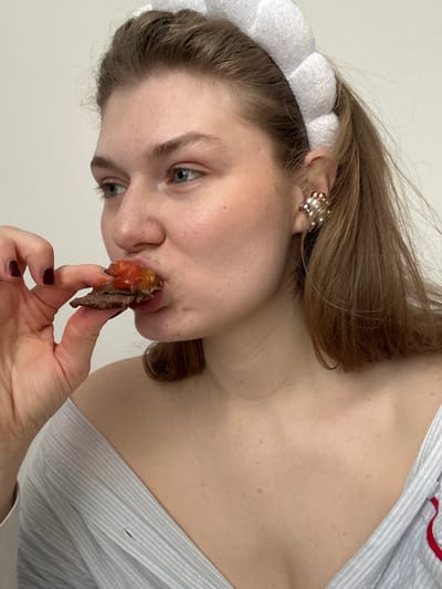 The scene, located indoors in Matiushino, Russia, depicts a young adult female from a close perspective. She has light brown hair and green eyes. She is wearing a white, segmented headband and a light-colored, off-the-shoulder garment with blue and white stripes; a red strap is visible on the garment's right side. A clustered earring featuring multiple pearls and gold-toned accents is present on her right ear. Her left hand, adorned with dark red nail polish, holds a small food item consisting of a dark, solid base topped with a reddish, semi-viscous substance. The subject is in the process of consuming this item, with her mouth partially open around it. The background consists of a plain, light-toned wall.