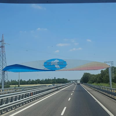 The image shows a highway scene with an overhead archway. The archway is white with a blue and orange-pink gradient on its underside. In the center of the archway is a blue oval with the text "45° PARALLELO" and a white outline of Italy. To the left of the archway, a tall, red and black lattice power pylon is visible. The highway itself is asphalt with white lane markings. Metal guardrails run along the sides of the road, with some orange markers on the guardrails. In the distance, there are trees and other vegetation, as well as some vehicles on the highway, including trucks and cars. The sky is clear and blue with a few white clouds. The scene appears to be a sunny day. The overhead archway indicates the location is near the 45th parallel, which passes through Voghera, Italy.