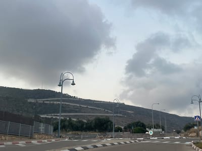 The image depicts a view of a winding road ascending a hillside in the city of Shlomi, Israel, under a cloudy sky. In the foreground, a paved road with black and white striped curbs is visible. Several streetlights with multiple lamps are positioned along the road. A metal fence runs along the left side of the road. The hillside is covered in vegetation and features a distinct series of switchbacks creating a winding path upwards. The sky is filled with dark, heavy clouds, suggesting overcast or potentially stormy weather. In the distance, on the very top of the hill, a tall tower or antenna structure is faintly visible. Traffic signs, including a pedestrian crossing sign, are present near the road.