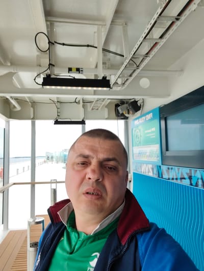 The image shows a man taking a selfie in what appears to be an indoor area with large windows looking out onto a bright, overcast day. The man is wearing a blue and red jacket over a green collared shirt. He has a neutral to slightly displeased expression.

The indoor setting features white ceilings with exposed electrical conduits and lighting fixtures. There are large windows that provide a view of an outdoor promenade or deck area with wooden flooring, metal railings, and some people in the distance. The sky outside is a uniform bright white, suggesting overcast weather.

To the right of the man, there is a bright blue wall with a wavy pattern and a digital display. Text is visible on this display, which reads "FIND YOUR WAYS" in large letters, with smaller text below that is difficult to decipher. There are also repeating blue text graphics with "WAYS" on the lower part of the blue wall.

The location context provided is Näset, Åland. This suggests the setting might be a ferry terminal, a cruise ship, or a similar waterfront facility in that region. The overall impression is of a man indoors, perhaps waiting or traveling, on a day with poor visibility.