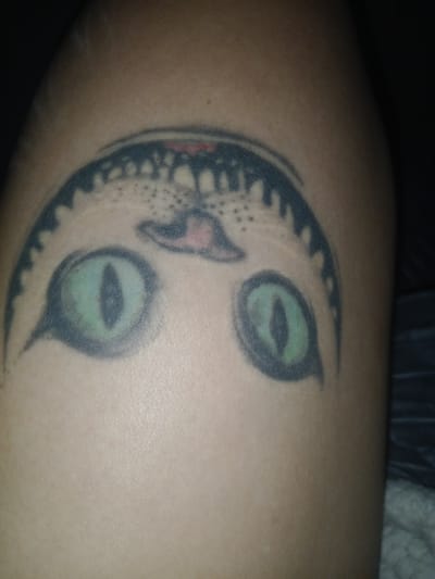 A close-up image shows a tattoo of a cat's face on a person's arm or leg. The tattoo is somewhat faded and appears to depict a stylized cat with large, green eyes, a pink nose, and black outlines. The location is not discernible.