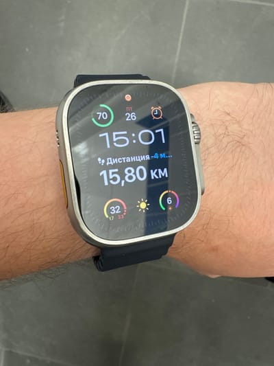 A person's wrist is shown with an Apple Watch Ultra on it. The watch face displays the time as 15:01, with a distance of 15.80 km. Other metrics visible include a heart rate of 70 bpm, a step count of 26, and a weather icon indicating sunny conditions with a temperature of 32 degrees. There is also an alarm icon and a rainbow ring with the number 6, possibly representing activity goals. The watch band is black. The background appears to be a grey, tiled surface, suggesting an indoor or urban outdoor setting. The person's arm is covered in hair. The scene is static, focusing on the watch face and the device on the wrist.
