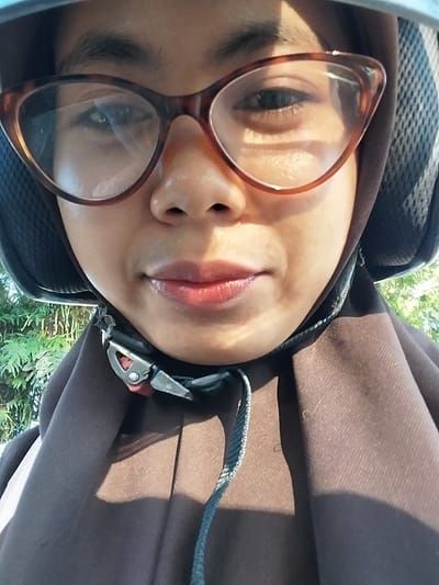The image is a close-up, selfie-style photograph of a person wearing a brown hijab and a helmet. The person has on glasses with brown frames. The focus is on their face, specifically their eyes, nose, and mouth. The background is out of focus, showing some greenery, possibly trees. The lighting suggests it is daytime, with sunlight casting shadows. The image appears to be taken outdoors, potentially in a humid environment given the slight sheen on the skin. There is no indication of the specific location within Macege, Indonesia. The photograph captures a portion of the individual's face and headgear.