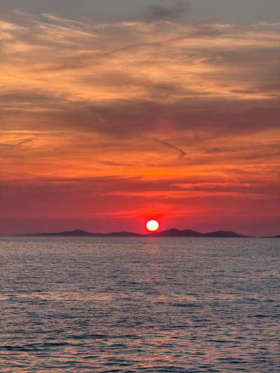 A vertical, eye-level shot captures a vibrant sunset over the Adriatic Sea, with the silhouette of islands in the distance, possibly near Primošten, Croatia. The sky is ablaze with fiery oranges and reds, transitioning to softer pinks and purples higher up, with wispy clouds streaked across. The sun, a brilliant white orb tinged with red, sits low on the horizon, casting a shimmering reflection on the rippling water below. The sea itself is a deep blue, with subtle hints of the sunset's colors reflecting on its surface. The overall impression is one of serene natural beauty at the end of the day.